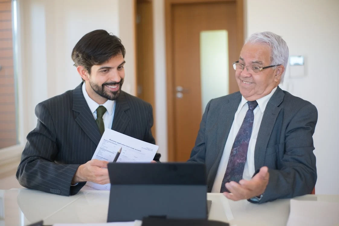 Two men wearing suits and tie in a conference room discussing to each other with a laptop on the table in front of them